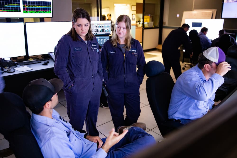 Two female employees talk to another employee seated at a computer desk